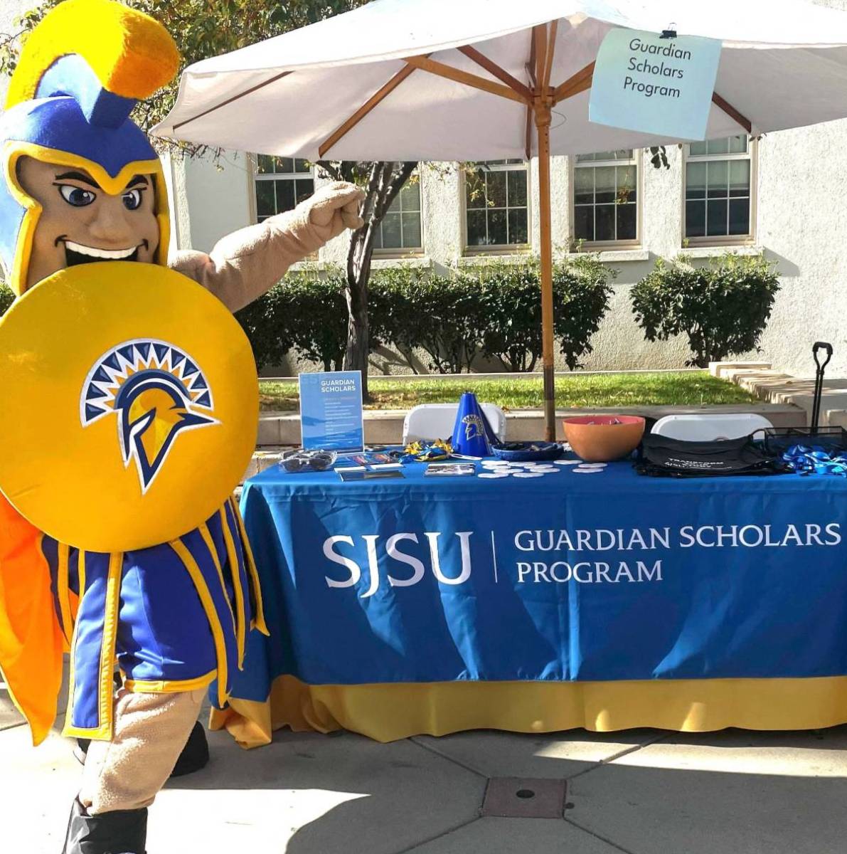 Mascot Sammy Spartan presenting a table with a Guardian Scholars banner
