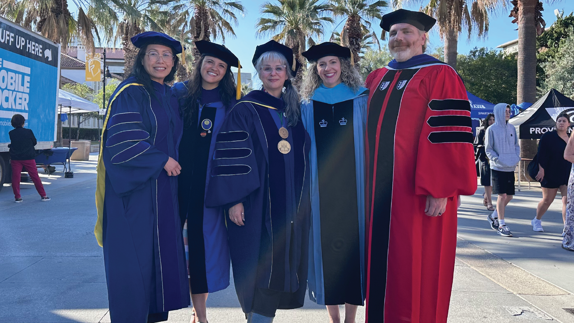 Five professors in their full doctoral regalia stand on the 7th Street Paseo before entering the event center for Spring 2025 Graduation.