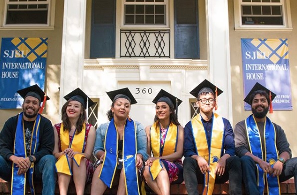 Students in graduation caps and sashes, seated on the brick porch of the International House