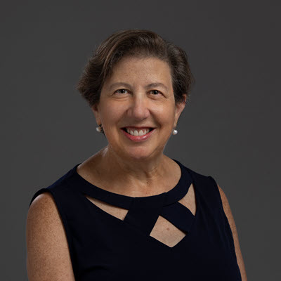 Portrait of Ruth Rosenblum. She a smiling, middle-aged woman with short brown hair, wearing a sleeveless navy top with a geometric cutout design. She is standing in front of a plain dark gray background.
