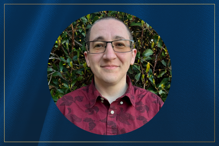 A person with glasses smiling for the camera wearing a burgundy colored shirt poses in front of a bush.