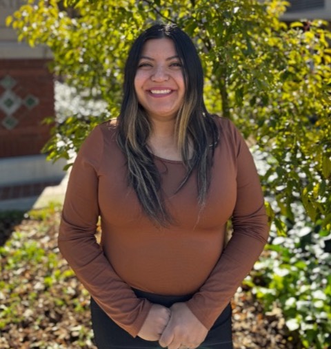 Hispanic woman with long, brown and blonde hair, wearing a gray cardigan and black shirt, smiling in a parking lot.