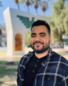 A man with short dark hair and a beard smiles at the camera while standing outdoors in a sunny area on the San José State University campus near the Arch of Dignity, Equality, and Justice. He’s wearing a blue and black plaid shirt over a black button-up shirt.
